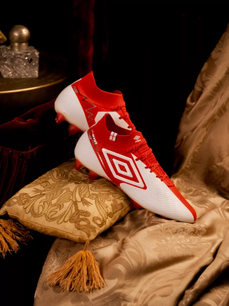 Par de chuteiras de campo Umbro brancas e vermelhas sobre uma almofada dourada