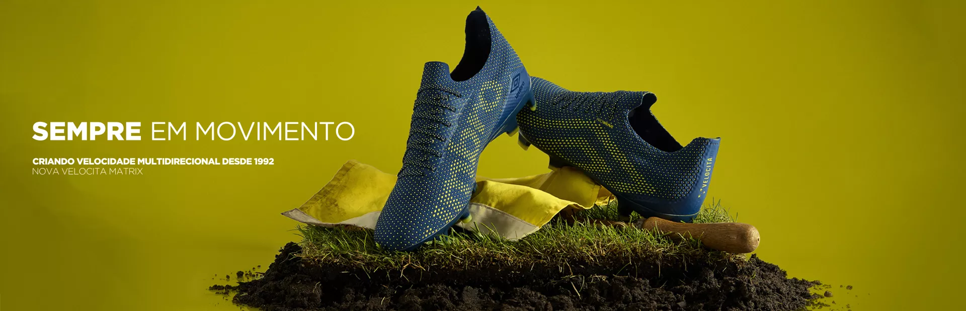 Chuteiras Umbro Velocità azuis sobre um torrão de grama, com o texto Sempre em movimento